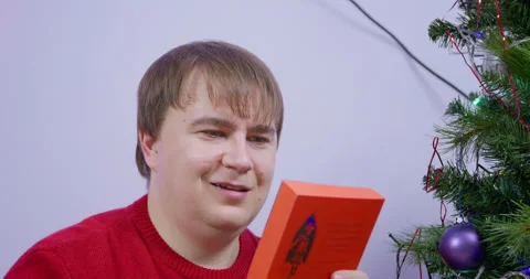 Young man in red knitted sweater unwraps his Christmas gift - a paperback book Stock Footage 220899751