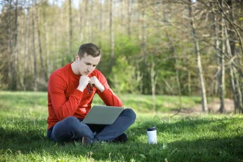 Young man in a red pullover with coffee cup and laptop working outside in a park Stock Photos