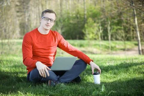 Young man in a red pullover with coffee cup and laptop working outside in a park Stock Photos