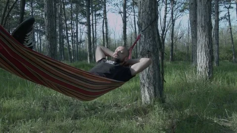 A young man relaxes on a hammock in the forest. Parzen with a beard swings on a Stock Footage