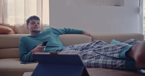 Young Man Relaxes on Sofa, Watching Television and Smiling in Pajamas on Weekend Stock Footage 306805214