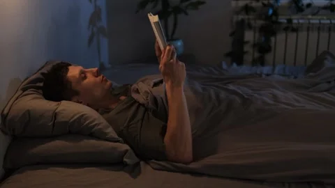 A young man relaxing in bed with soft evening lighting, reading a book and embra Vídeo Stock 296424535