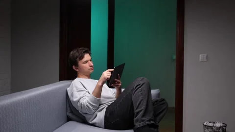 Young man relaxing on a comfortable sofa in a dimly lit room, focusing on his Stock Footage 346251326