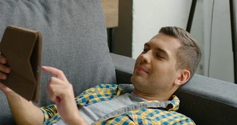 Young man is relaxing on a couch and playing game on a tablet Stock Footage 112763838