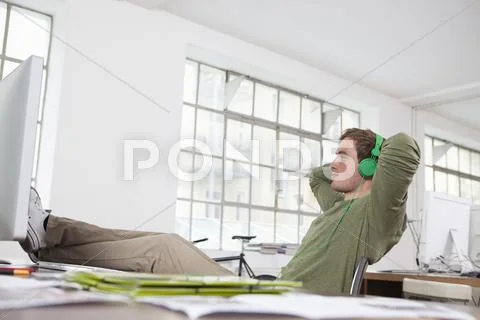 Young Man Relaxing With Feet On His Desk And Hearing Music With Headphones In