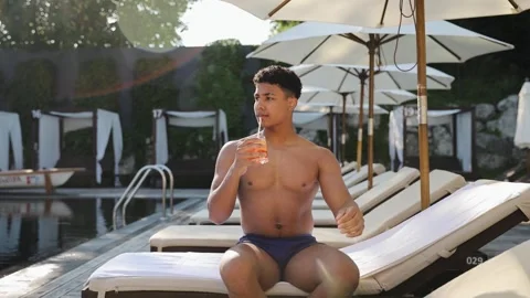 Young man relaxing by hotel pool, enjoying refreshing drink on a lounge chair Stock Footage 333564958