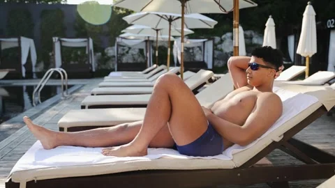 Young man relaxing by the pool, sunbathing on a lounge chair at luxury resort Stock Footage 328160176