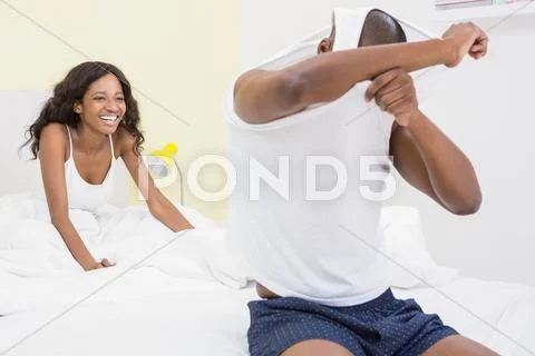 Young man removing his vest sitting on the bed Stock Photos