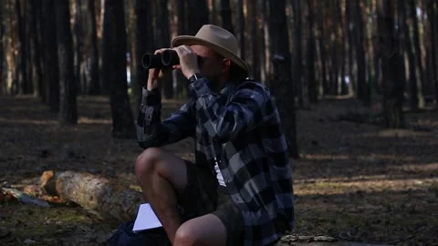 Young man resting on a log and looking through binoculars while hiking Stock Footage 150255608