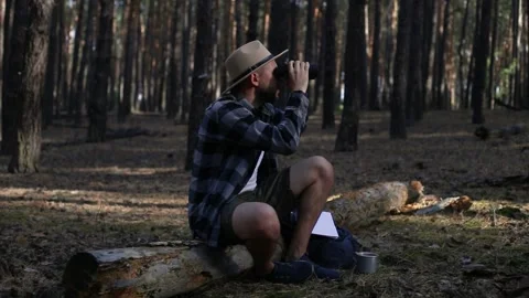 Young man resting on a log and looking through binoculars while hiking Stock Footage 150255692