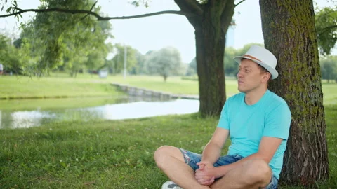 Young man is resting while sitting under a tree in the park, tracking by camera Stock Footage 138999781
