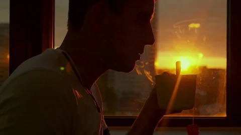 A young man rests in the evening at sunset and drinks hot tea by the window. HD Stock Footage 81714773