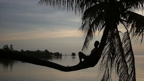 A young man rests sitting on a palm tree, above the water against the backdrop Stock Footage 95940322