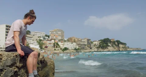 Young Man on Rock at Windy Beach Seaside in Sunshine. Long Bun Hair Hipster  Stock Footage 94429329