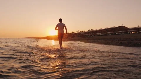 Young man running on beach at sunset toward golden sun over sea horizon. Stock Footage 334586598