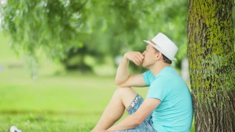 Young man is sad while sitting under a tree in a city park, blurred background Stock Footage 148039060