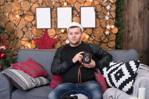 Young man in a Santa Claus hat sits on the couch with a camera Stock Photos