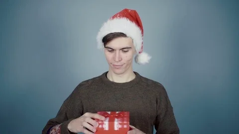 Young man in Santa hat opening a gift box with a surprise inside seeing funny Stock Footage 70223415