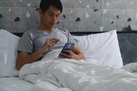Young man scanning QR code on smartphone while relaxing in bed 写真素材