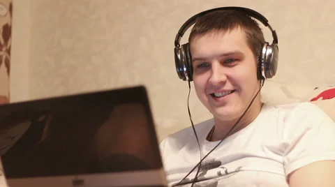 Young man seated on couch working with a laptop and listen music in headphones Stock Footage 61170455