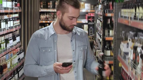 Young man selecting beer while checking his phone in a vibrant supermarket aisle Stock Footage 311895606