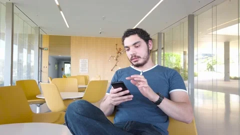 Young man sends a voice message with his mobile, sitting in lobby of office Stock Footage 136924019