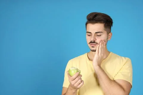 Young man with sensitive teeth and apple on color background. Space for text Stock Photos