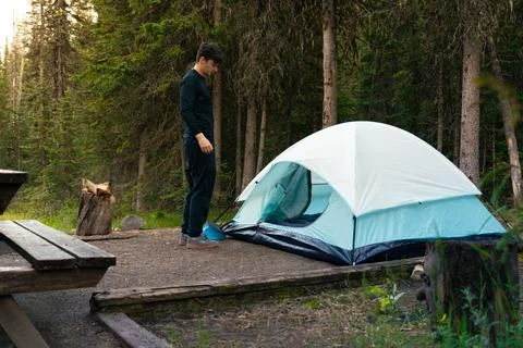 Young man sets up a white and blue tent in a forest campsite alone. Stock Photos