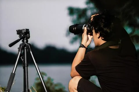 Young man setting up camera on the edge of the lake. Back view of male Sett.. Stock Photos