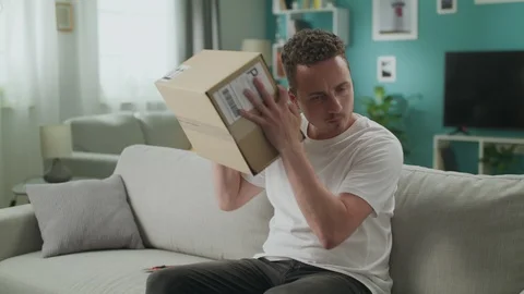 Young man shakes and opens cardboard parcel with interest 動画素材 108548987