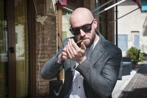 Young man with a shaved head, beard, and sunglasses, lights up a cigar. Stock Photos