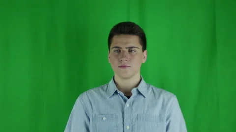 Young man shifts in his seat in front of a greenscreen in a blue shirt Vidéo 59084802