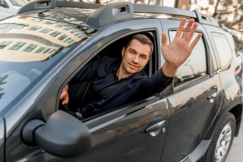 A young man in a shirt sits in his car and waves to someone. Taxi driver looks Stock Photos