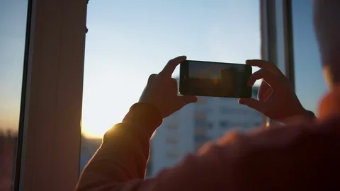 A young man is shooting a sunset on a mobile phone through a window. Stock Footage 130157673