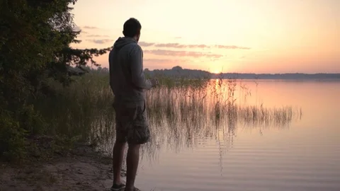 The young man in shorts drinks tea at dawn on lake. Stock Footage 79392824
