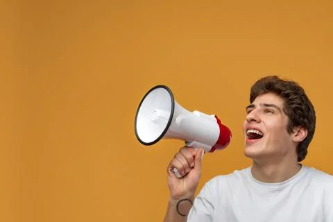 Young man shouting into megaphone making announcement against yellow background Stock Photos