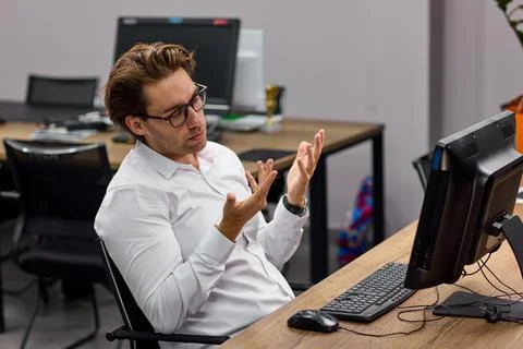 Young man shrugging beside computer in empty office, showing frustration and Stock Photos