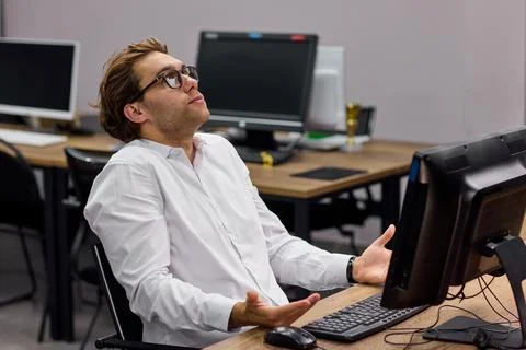Young man shrugging beside computer in empty office, showing frustration and Stock Photos