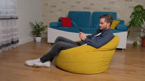 Young man sits on bean bag and playing games on phone, at home. Stock Footage 140910185