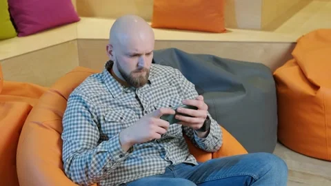 Young man sits on a bean bag while playing online using his phone. Video stock 151546844