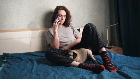 A young man sits in bed and calls someone, the dog sleeps between his legs. Stockbeeldmateriaal 196813193
