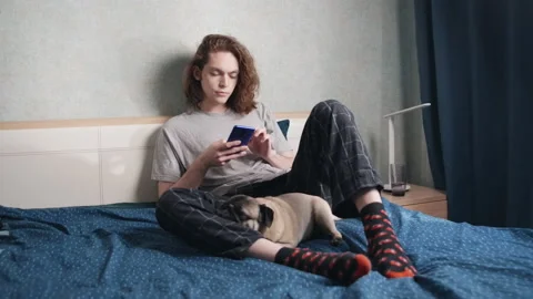 A young man sits in bed and looks on the phone, a dog lies and sleeps nearby. 動画素材 196813240