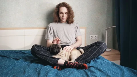A young man sits in a bed on top of him is a dog. The guy strokes the dog 動画素材 196813373