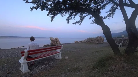 Young man sits on the bench on the beach near the tree Stock Footage 114222025