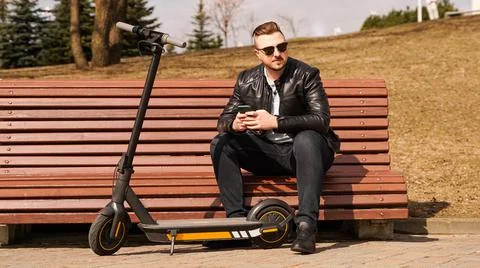 Young man sits on a bench on a spring day. Electric scooter next to a bench Stock Photos