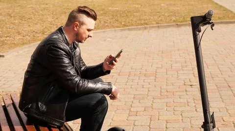Young man sits on a bench on a spring day. Electric scooter next to a bench Stock Photos