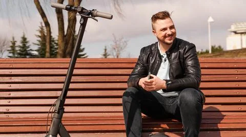 Young man sits on a bench on a spring day. Electric scooter next to a bench Stock Photos