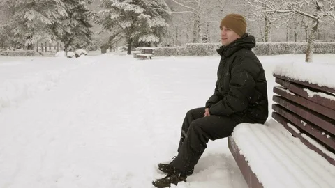 A young man sits on a bench in winter park and admiring the snow. A man in a Stock Footage 71221333