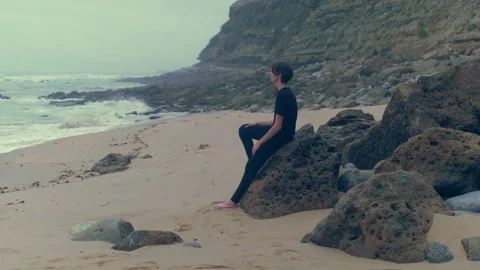 Young man sits on a  big stone on the empty seashore, cloudy rainy weather... Stock Footage 261804791