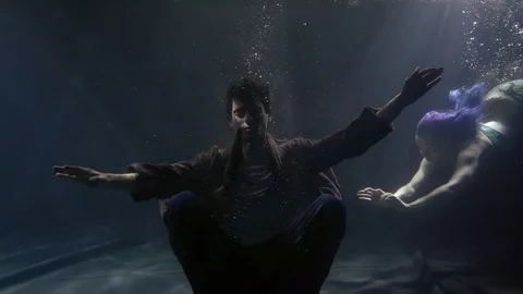 A young man sits at the bottom of a dark ocean and a beautiful mermaid with blue 스톡 동영상 105129404